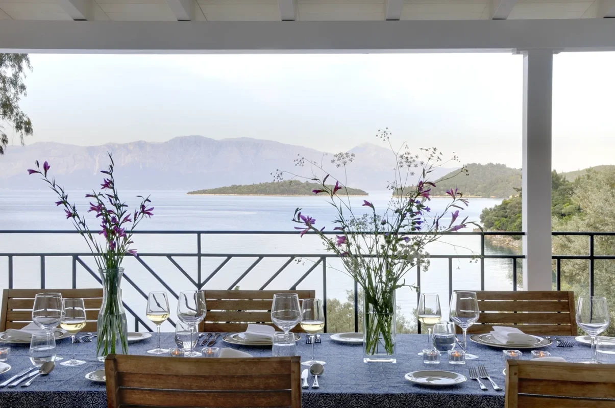 legant outdoor dining setup on a terrace overlooking the sea and mountains, decorated with glassware and fresh flowers.