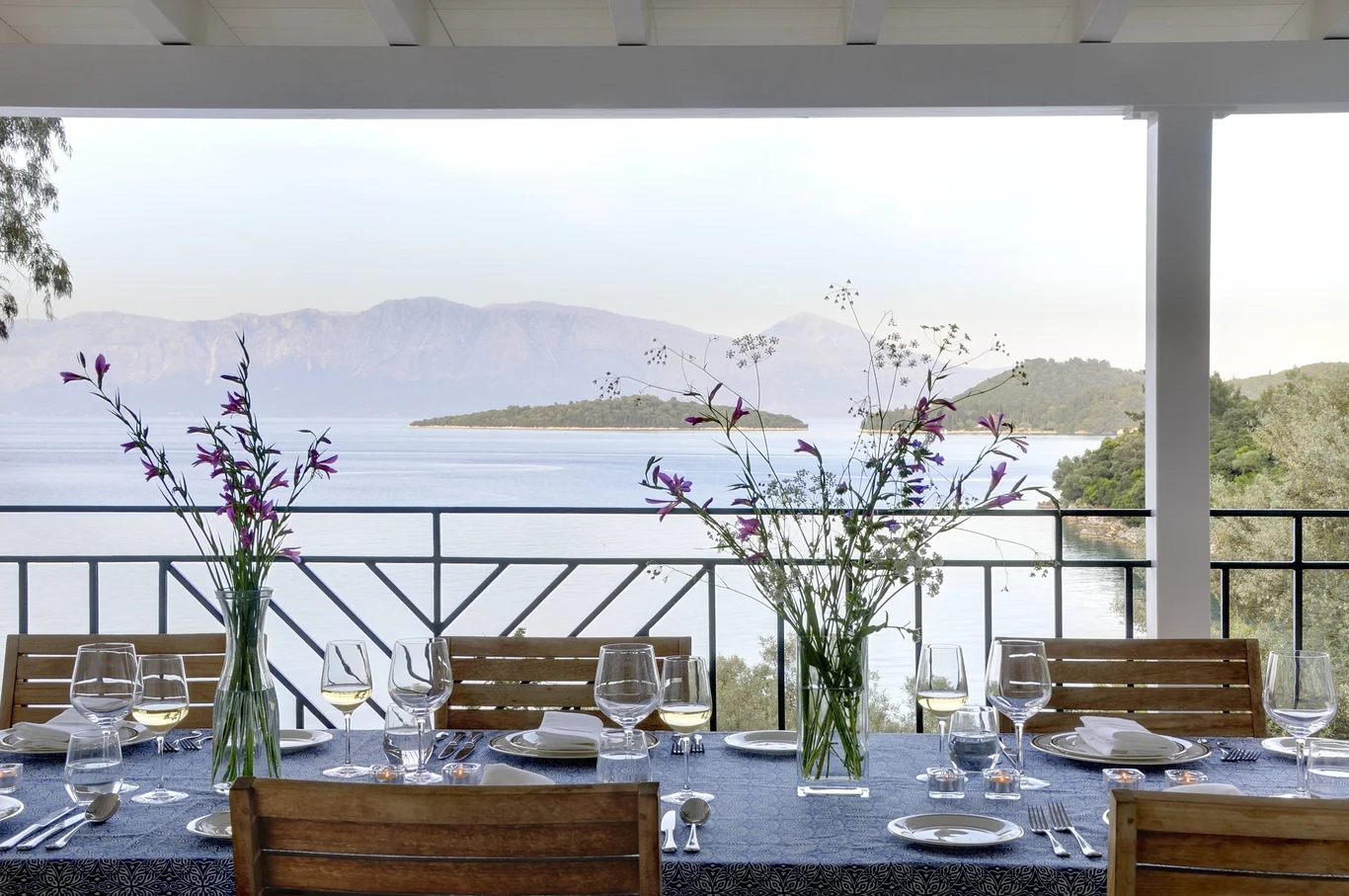legant outdoor dining setup on a terrace overlooking the sea and mountains, decorated with glassware and fresh flowers.