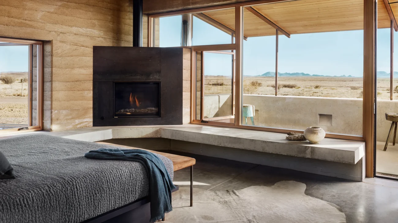 Rustic modern bedroom with desert views, rammed earth walls, and a fireplace framed by large windows.