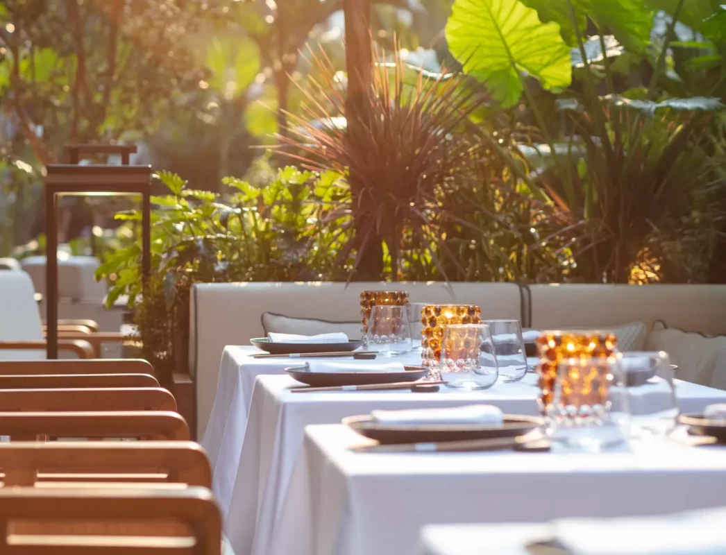 Refined restaurant terrace with white tablecloths, amber glass candle holders, and surrounding tropical greenery at sunset.