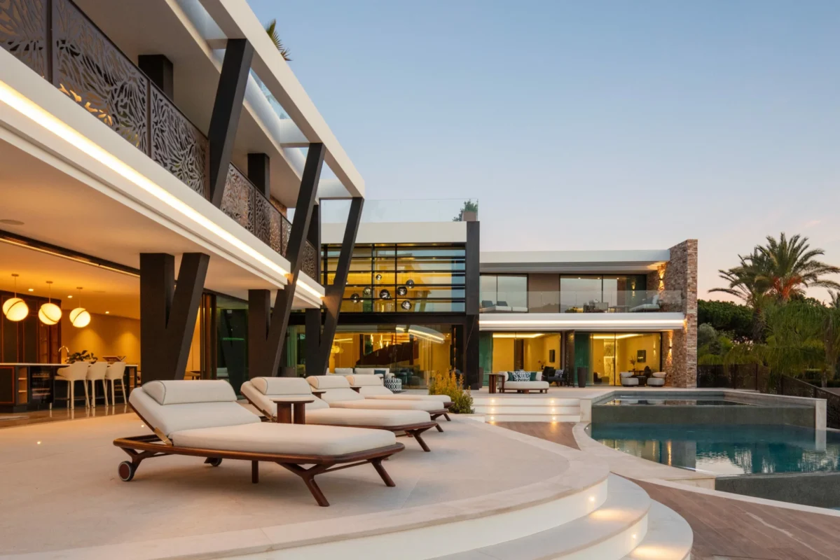 Luxury villa exterior with modern architecture, poolside loungers, and an illuminated infinity pool at sunset.