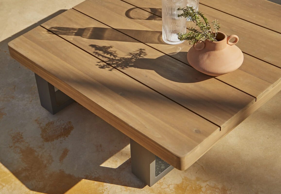 Close-up of a modern wooden outdoor coffee table with terracotta vase and glass carafe in natural sunlight.