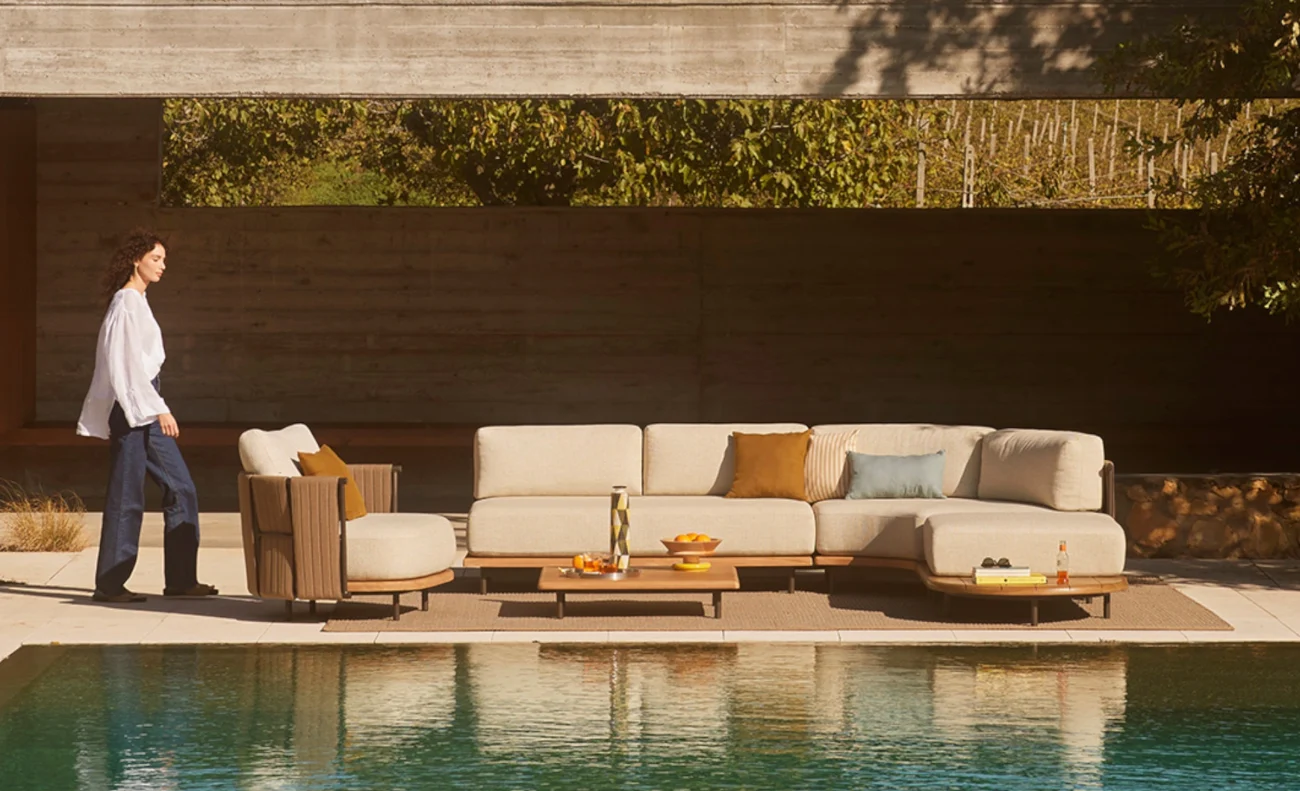 Modern outdoor poolside lounge with beige sectional sofa, armchair, and low wooden tables styled with colorful cushions