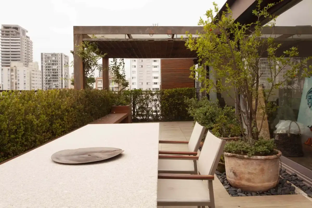 Minimalist rooftop dining area with stone table, wooden pergola, and greenery overlooking city skyscrapers.