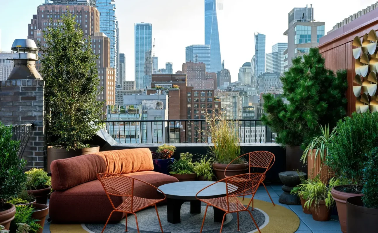 Urban rooftop lounge with rust-colored sofa, metal chairs, and lush greenery set against a New York City skyline backdrop.