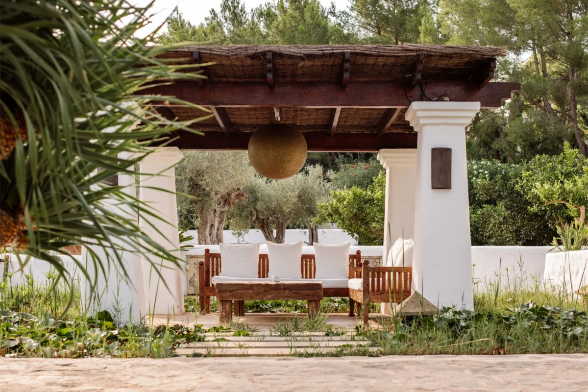 Serene Ibiza garden lounge with wooden benches, white cushions, and shaded pergola surrounded by olive trees.