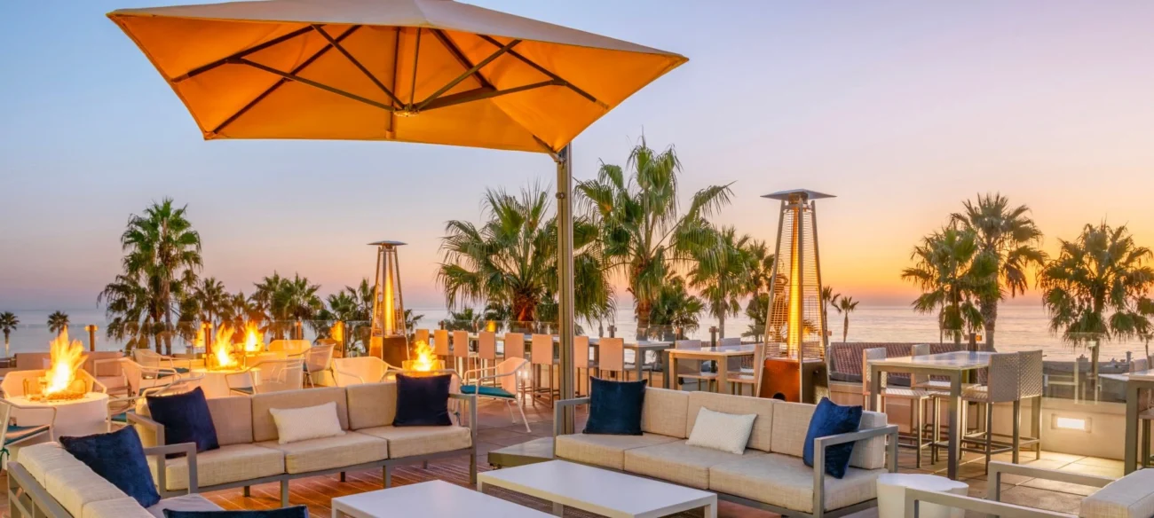 Luxury outdoor lounge furniture arranged on a seaside terrace at sunset, featuring elegant outdoor seating, fire tables, and palm-lined coastal views.