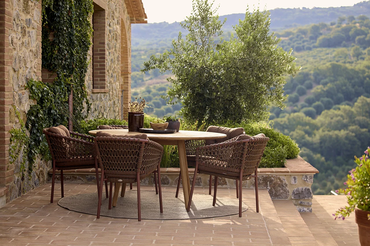 Luxury outdoor dining set with woven rope chairs and a wooden table on a Mediterranean stone terrace overlooking rolling green hills.