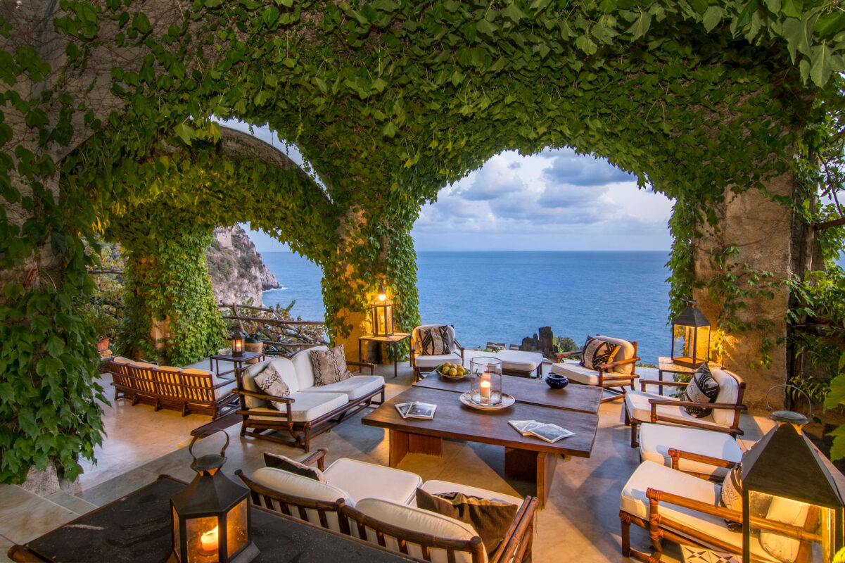 Luxury outdoor lounge beneath vine-covered stone arches overlooking the Amalfi Coast, featuring Mediterranean seating and lantern lighting