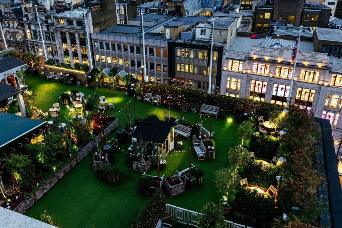 Evening view of a landscaped rooftop garden in central London featuring layered planting, ambient lighting, and multiple outdoor seating zones.