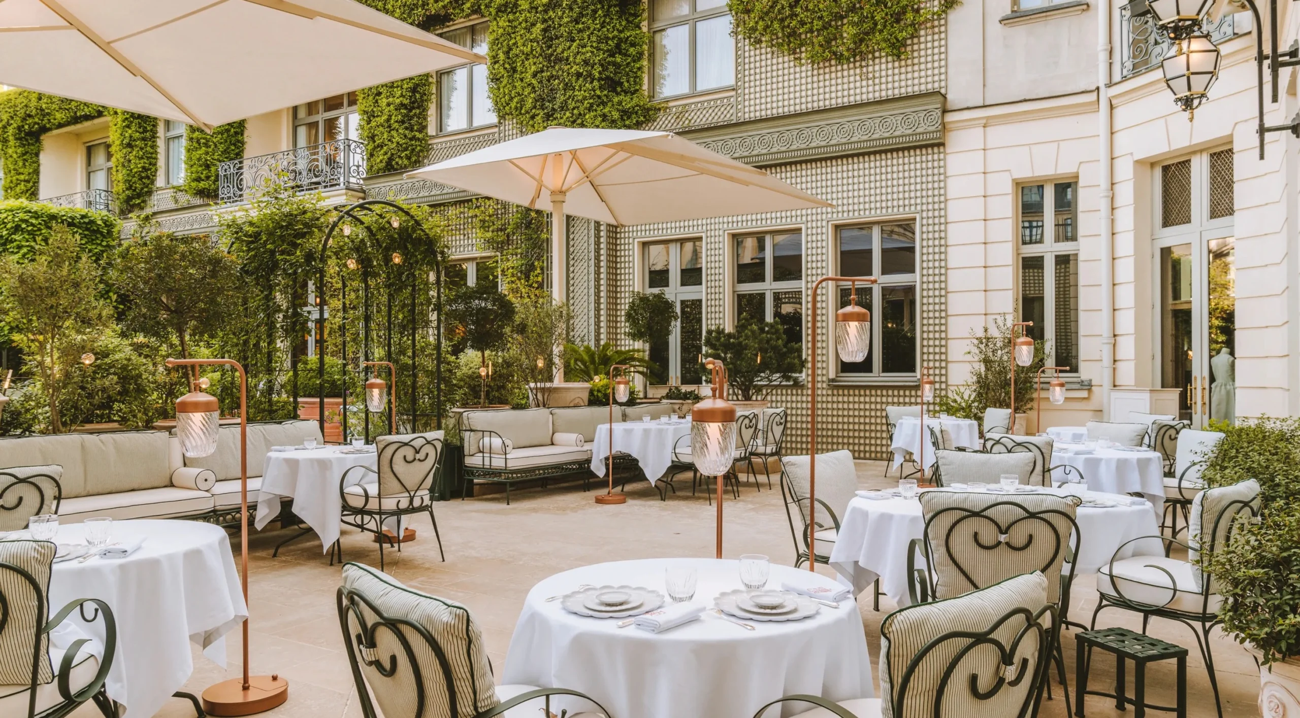 Charming Paris courtyard terrace with white tablecloth café tables, wrought-iron chairs, large umbrellas, greenery-covered walls, and classic architectural details.
