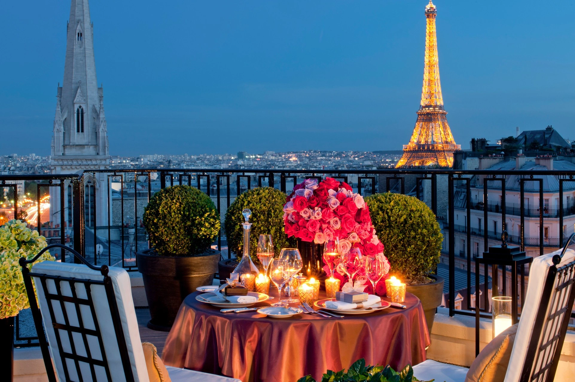 Romantic rooftop dining setup in Paris with a candlelit table, pink rose centerpiece, wine glasses, and the illuminated Eiffel Tower at dusk.