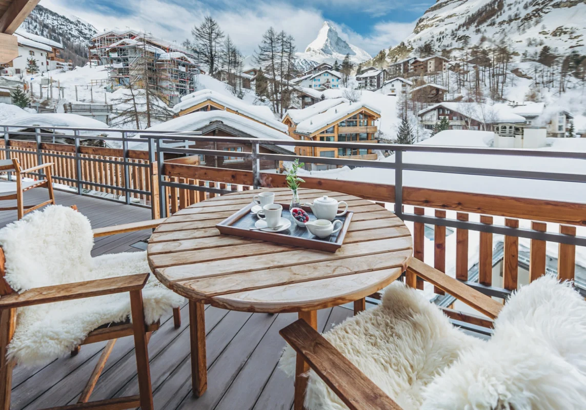 Luxury Swiss mountain chalet balcony with wooden outdoor dining furniture, sheepskin chairs, and panoramic winter views over snow-covered alpine village and peaks.