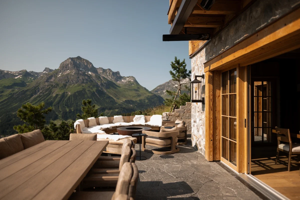 Contemporary alpine chalet terrace featuring refined outdoor lounge seating, natural stone flooring, and uninterrupted mountain views in a high-altitude summer setting.