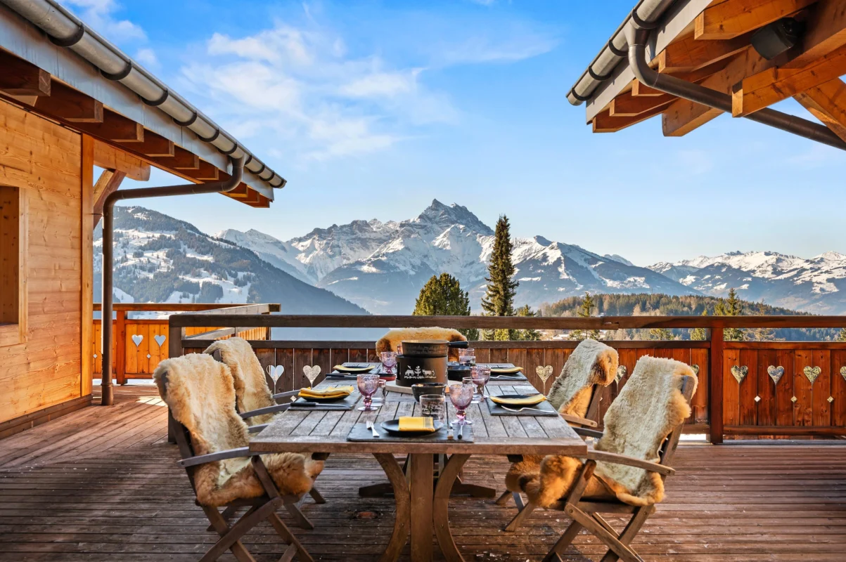 Traditional Swiss chalet outdoor dining terrace with wooden table and chairs, layered with sheepskin textiles, overlooking dramatic snow-capped Alpine mountains.