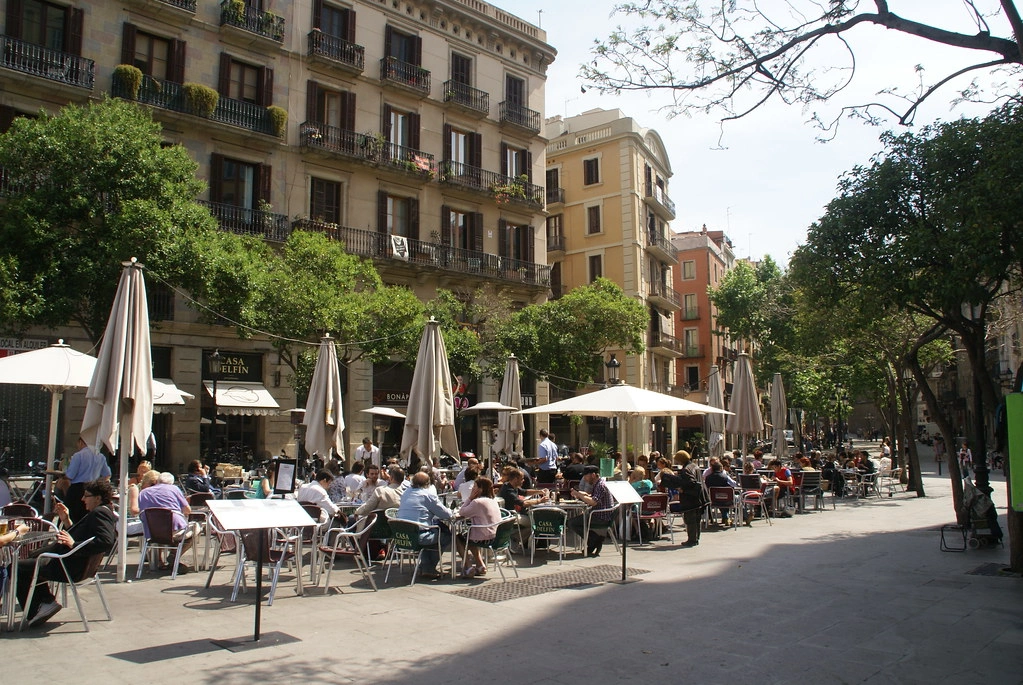Sunlit Barcelona plaza with outdoor café seating beneath trees, showcasing Mediterranean outdoor living design integrated into daily city life.