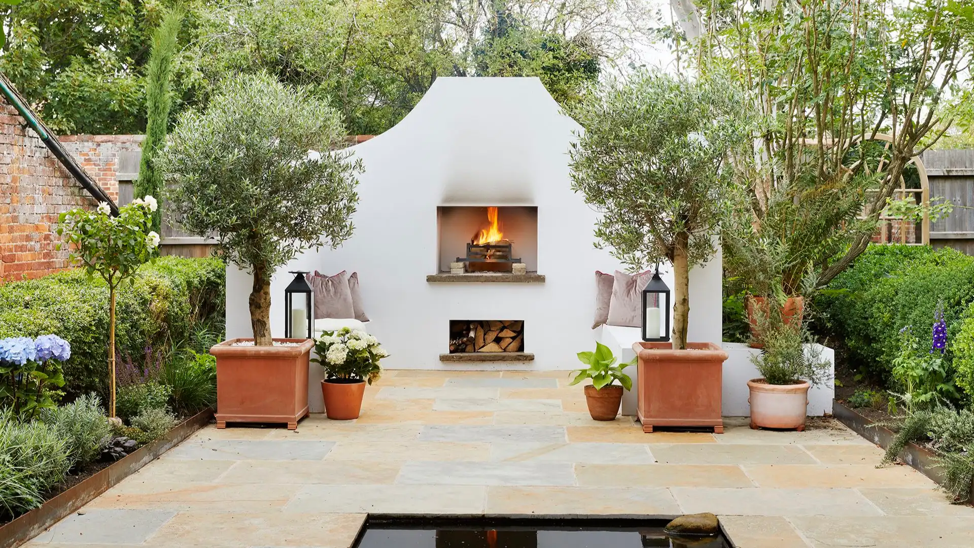 Sunlit Greek island courtyard with whitewashed walls, limestone flooring, pergola-cast shadows, and minimalist Mediterranean seating arranged around an olive tree overlooking the Aegean Sea.
