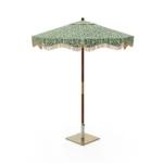 Matera parasol with a green ikat-patterned canopy and fringe stands on a sun-drenched terrace with stone walls, cacti, and potted plants