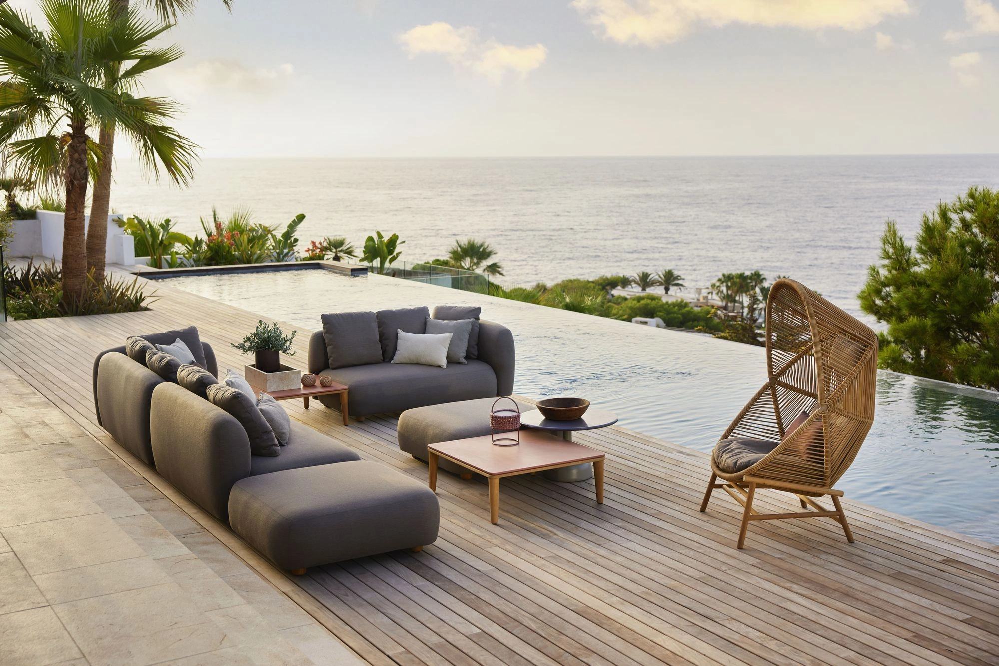 Luxury outdoor living terrace overlooking the sea, featuring contemporary outdoor furniture with modular sofas, woven lounge chair, and minimalist teak tables in a serene Mediterranean landscape.