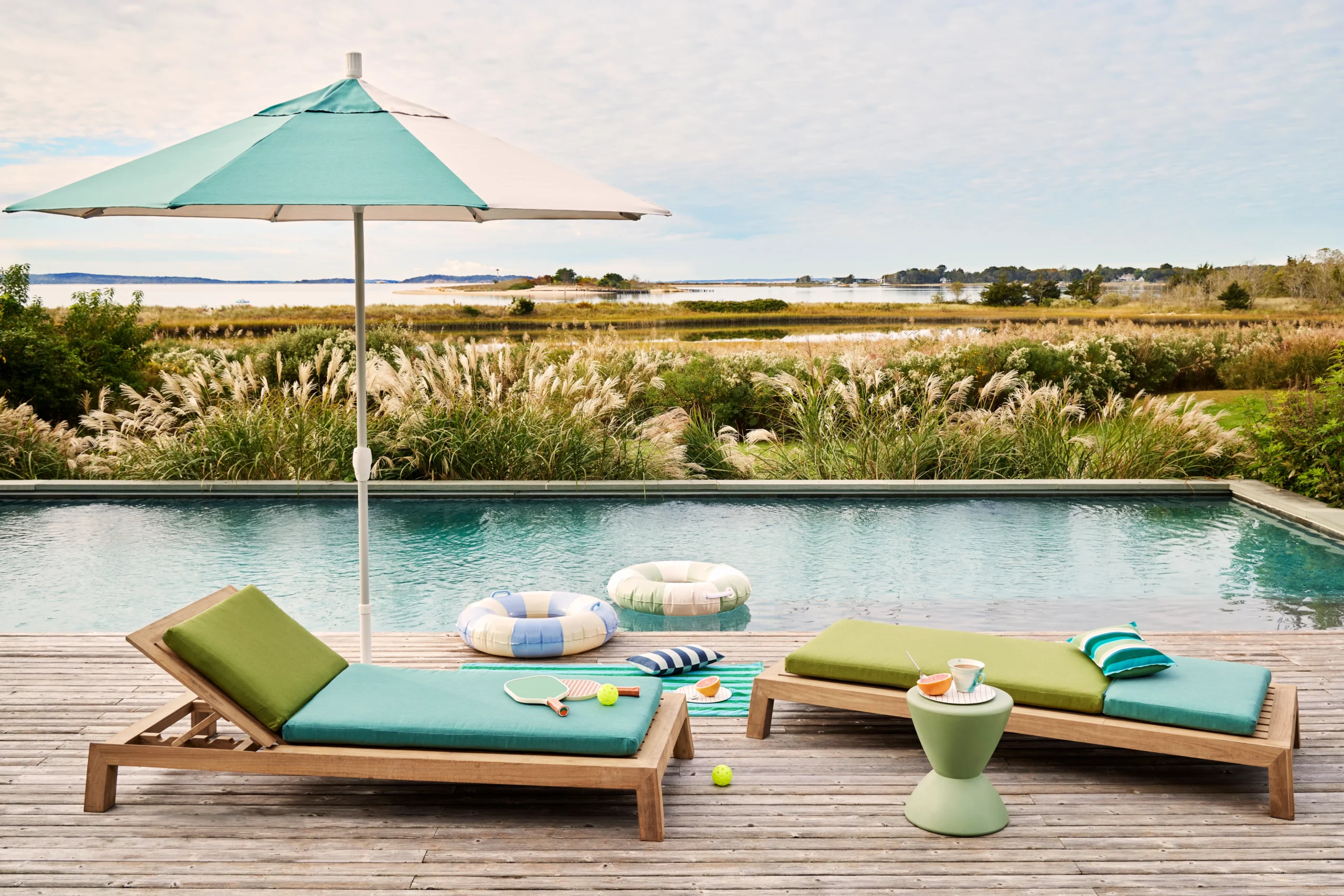 Luxury poolside furniture including teak poolside lounge chairs and upholstered daybeds arranged on refined terraces beside infinity pools, overlooking coastal and desert landscapes, designed for elegant outdoor living and resort-style comfort.