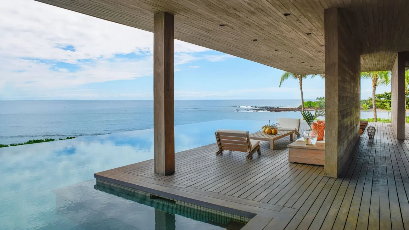 Luxury teak outdoor furniture including lounge seating, dining table, and sectional sofas styled on contemporary oceanfront terraces and poolside decks, showcasing natural wood tones and refined coastal architecture.