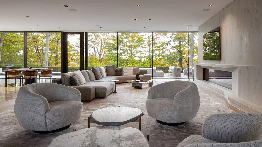 Luxurious modern living room with large glass windows showing a view of trees. Features gray sofas, a marble coffee table, and a sleek fireplace.