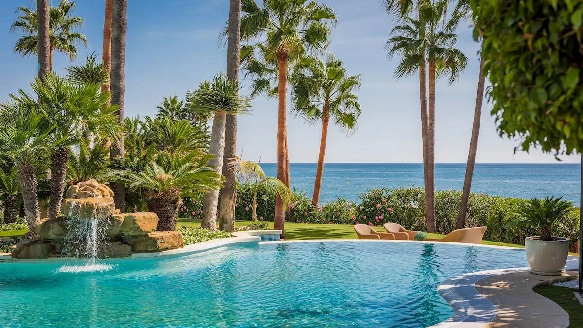 Tropical oasis with a curved pool, waterfall, and palm trees. Lounge chairs face the ocean under a clear blue sky, conveying tranquility.