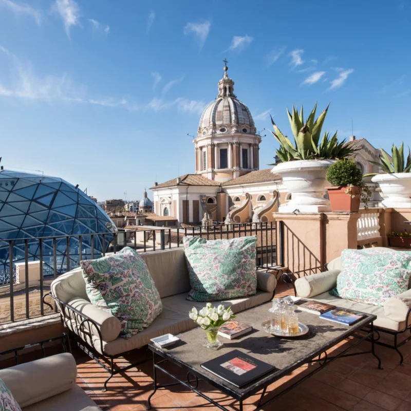 Rooftop terrace with beige cushioned seating, floral pillows, and a table with drinks and books. View of Rome's baroque domes under a clear sky.