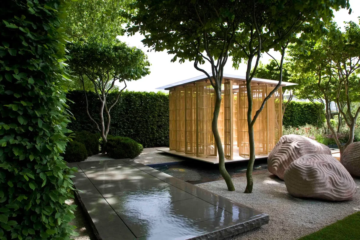 A serene garden features a wooden pergola surrounded by lush greenery. Smooth stone pathways and sculpted rocks add tranquility under shaded trees.