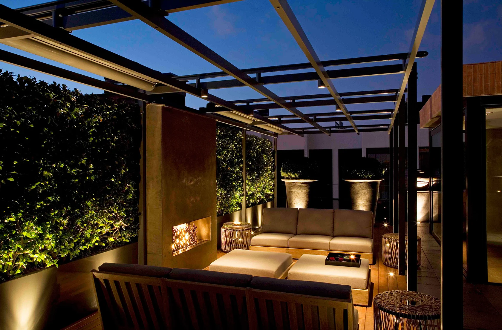 Rooftop lounge at dusk, cozy seating under a modern pergola. Soft lighting, lush greenery, and warm tones create an inviting, tranquil atmosphere.