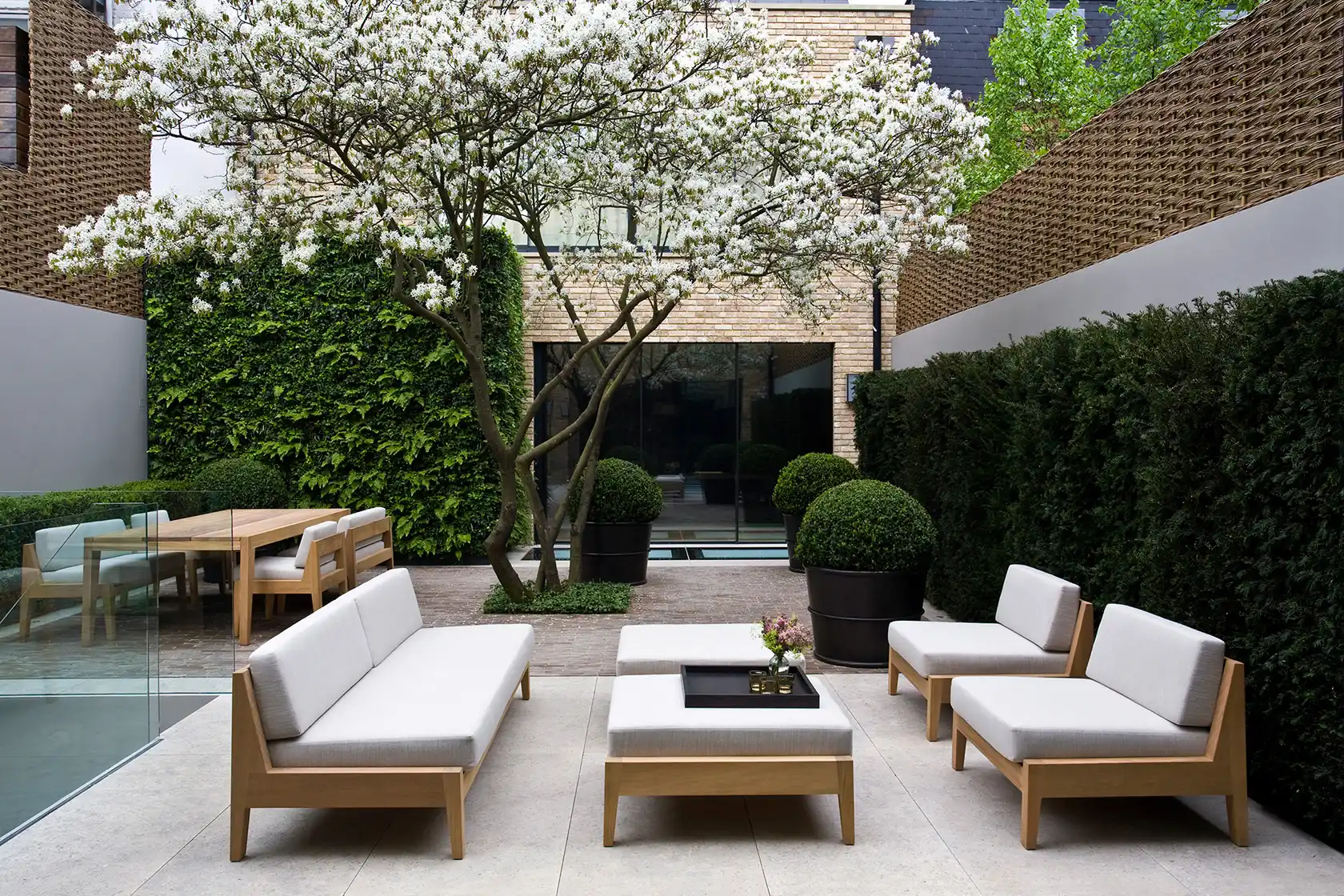 Modern courtyard with minimalist wooden furniture, blossoming tree, and lush green hedges. A peaceful and inviting outdoor space.
