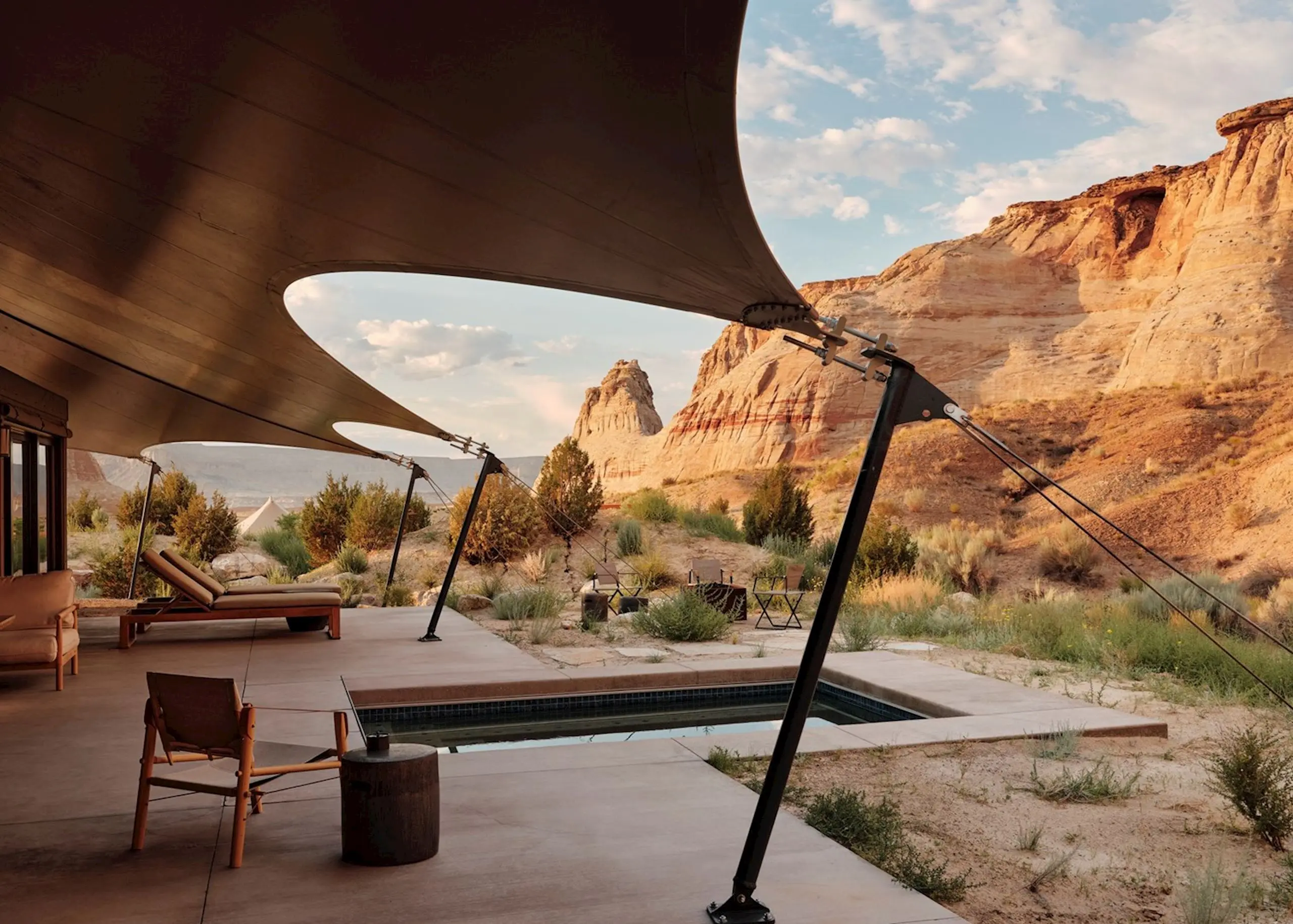 Luxurious patio with modern canopy, lounge chairs, and a small pool overlooks desert landscape and rocky cliffs under a partly cloudy blue sky. Relaxing ambiance.