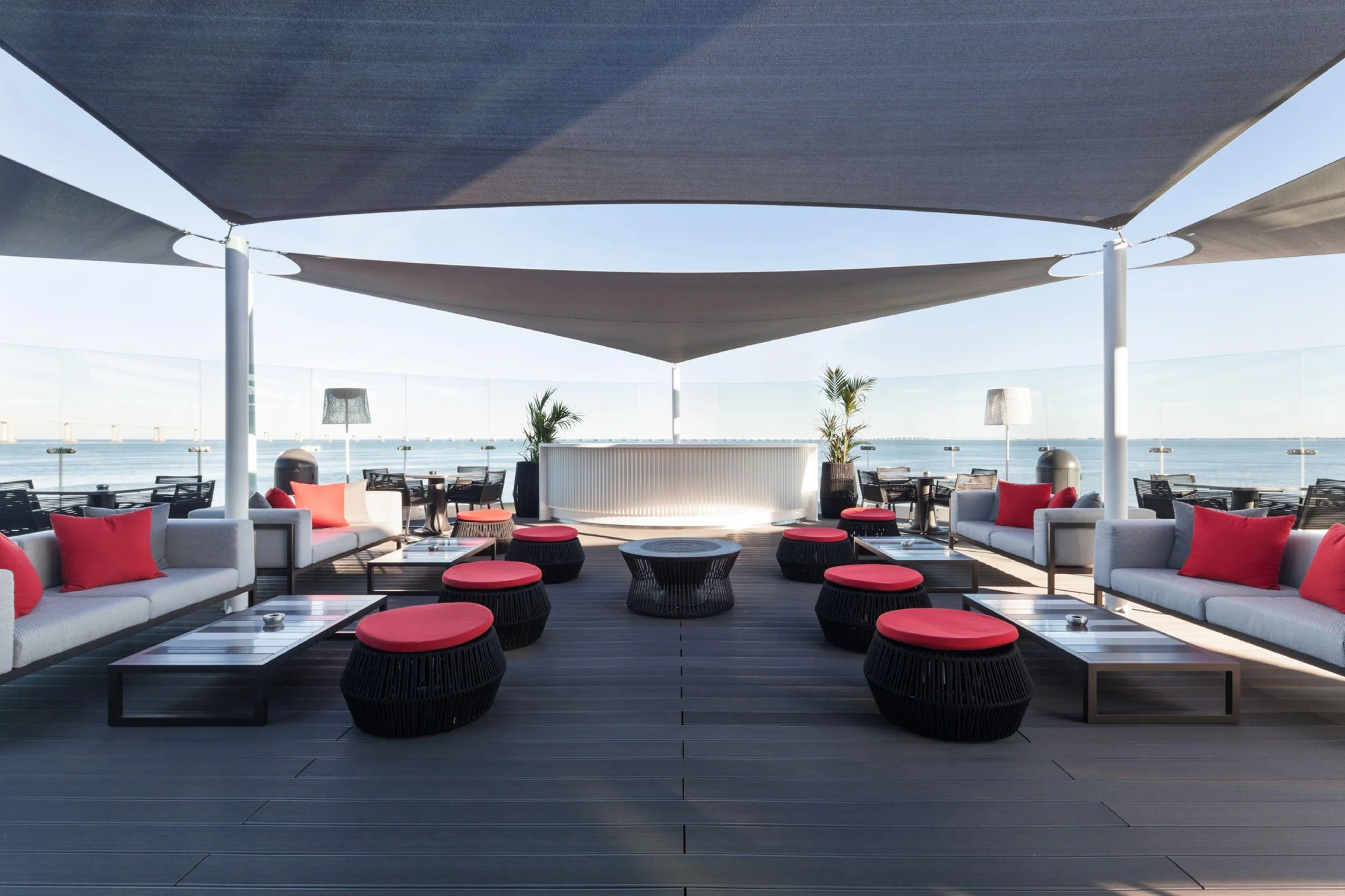 A modern rooftop terrace with sea views, featuring cozy sofas with red cushions, round tables, and overhead shade sails. The atmosphere is relaxing.
