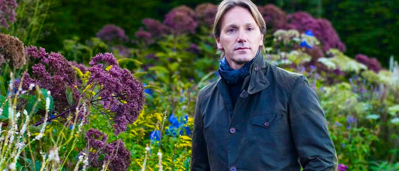 Luciano Giubbilei in a green jacket stands in a vibrant garden filled with purple, yellow, and blue flowers. The scene conveys a peaceful, natural ambiance.