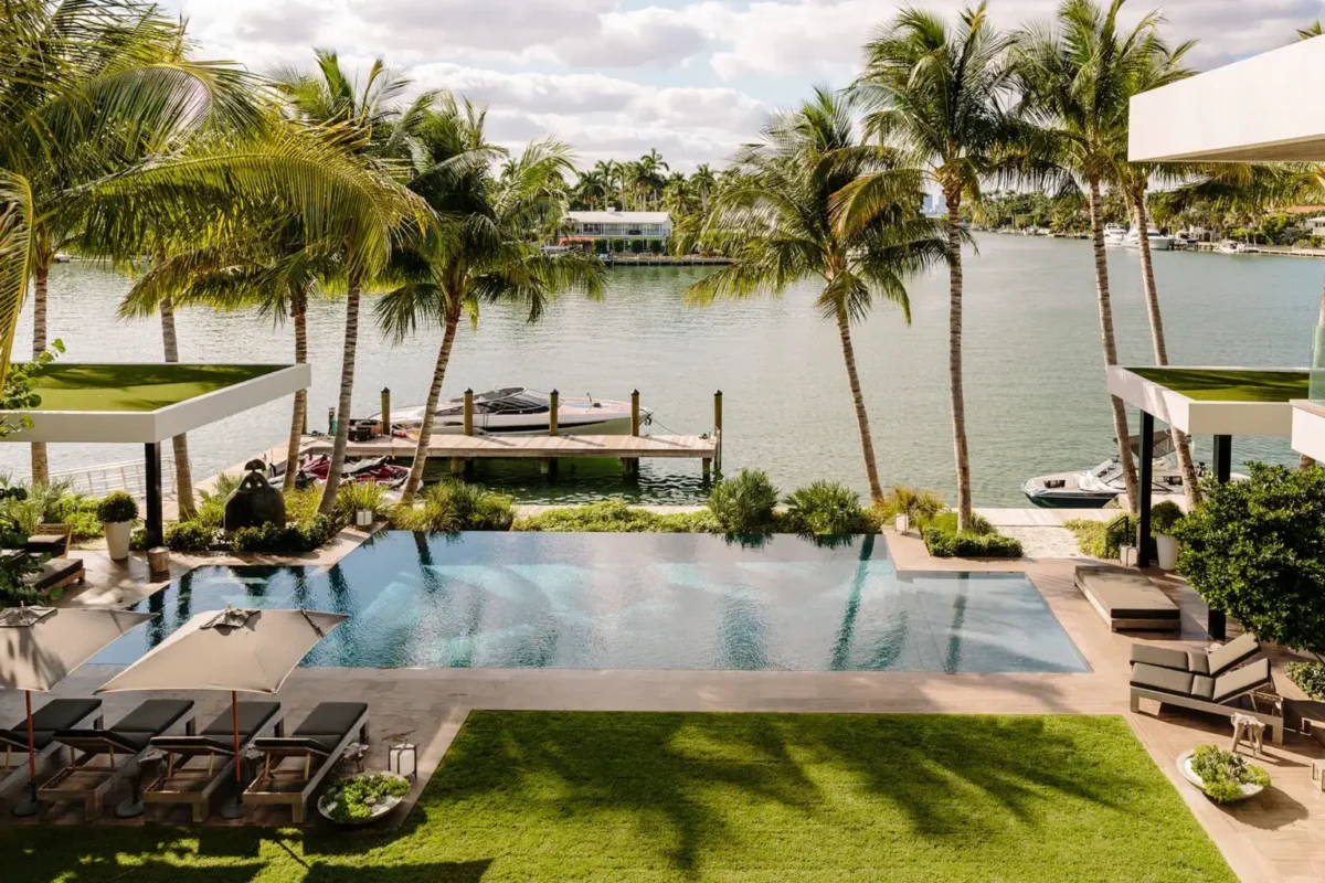 Luxurious waterfront scene with a pool surrounded by palm trees, sun loungers, and a dock set on a tranquil, sunny river. Relaxing and upscale atmosphere.