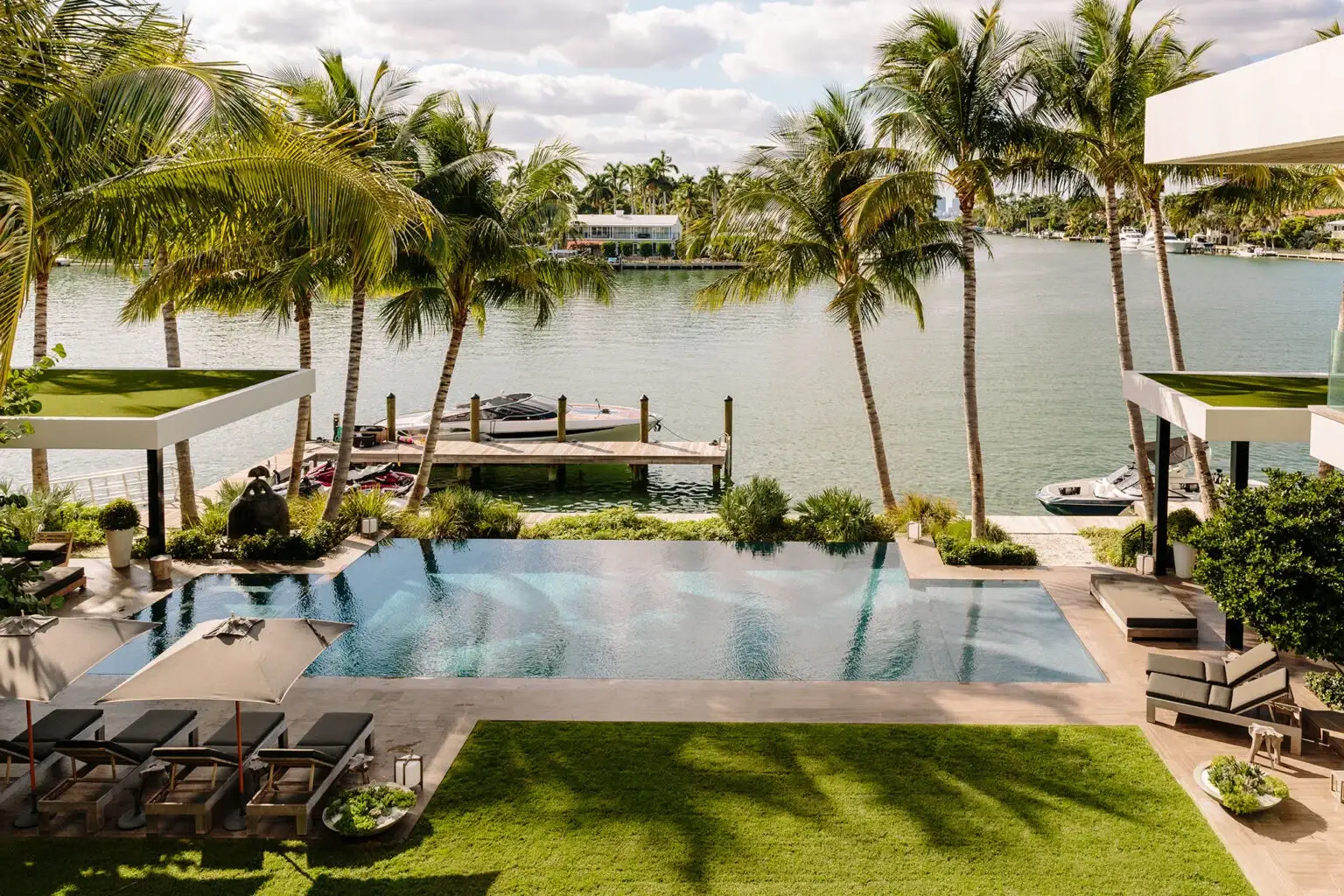 Luxurious waterfront scene with a pool surrounded by palm trees, sun loungers, and a dock set on a tranquil, sunny river. Relaxing and upscale atmosphere.