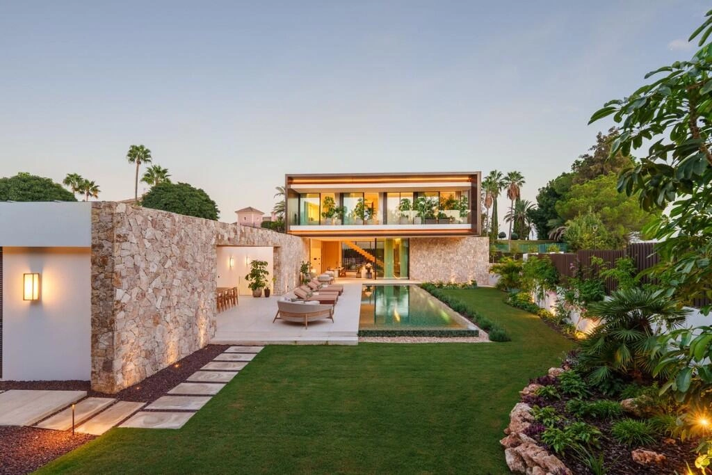 Modern villa with stone walls, large windows, and lush greenery. Features a reflective pool, patio seating, and expansive lawn under a clear sky.