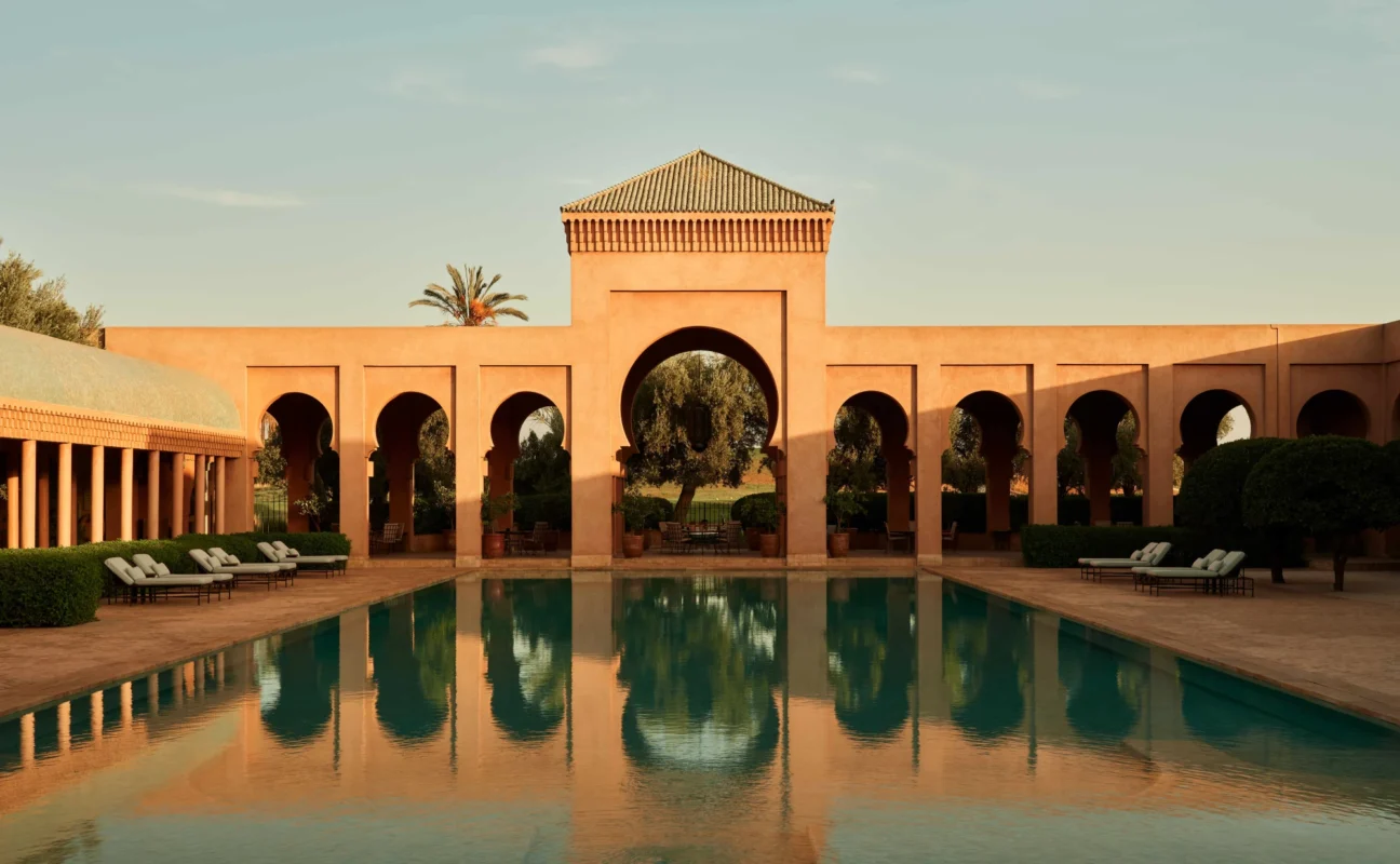 A serene pool reflects an elegant arched pavilion under a clear sky, flanked by lounge chairs and trees, evoking a sense of luxury and tranquility.