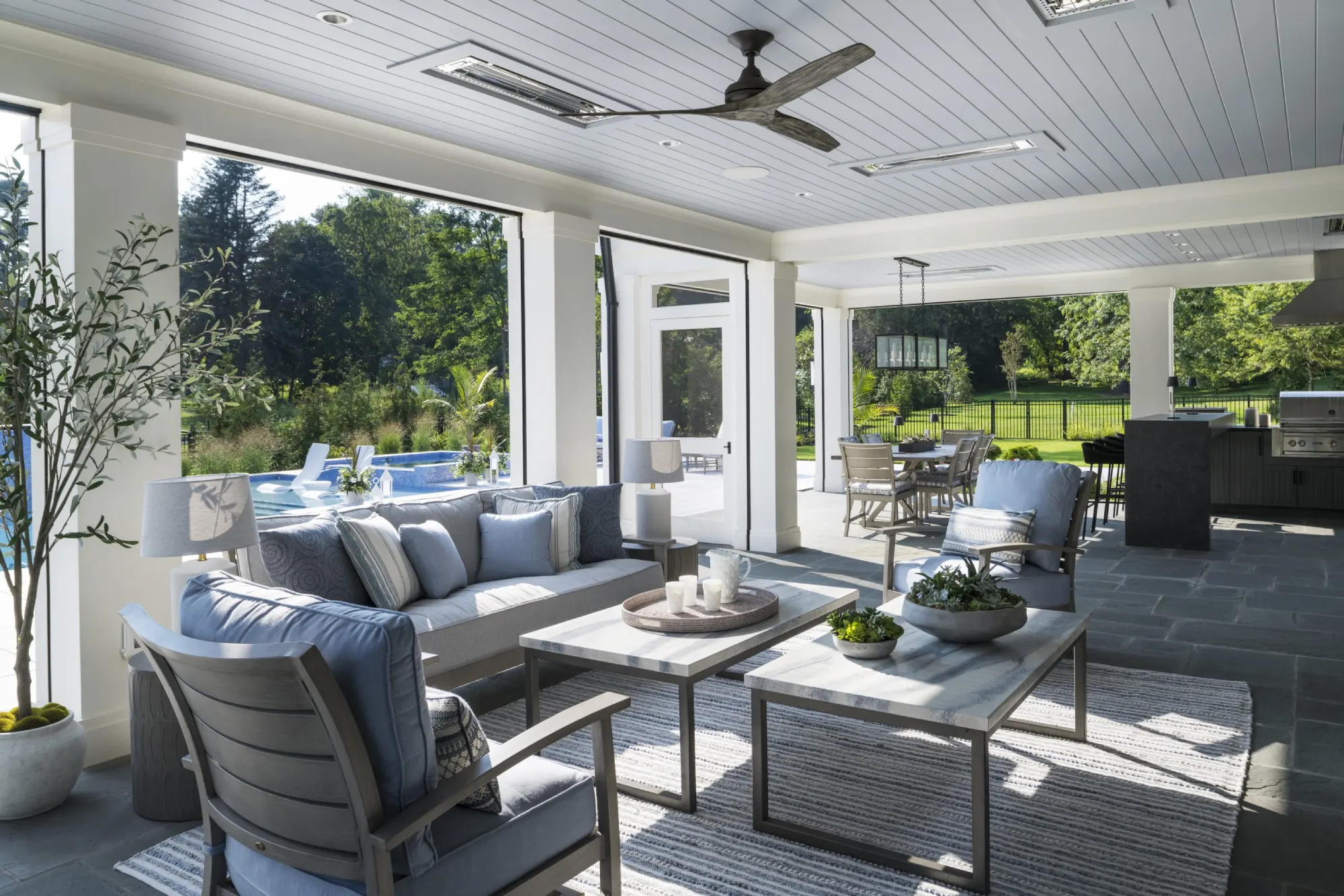 Modern outdoor patio with cozy seating, blue cushions, and coffee table on a striped rug. Overhead fan and lush greenery create a serene atmosphere.