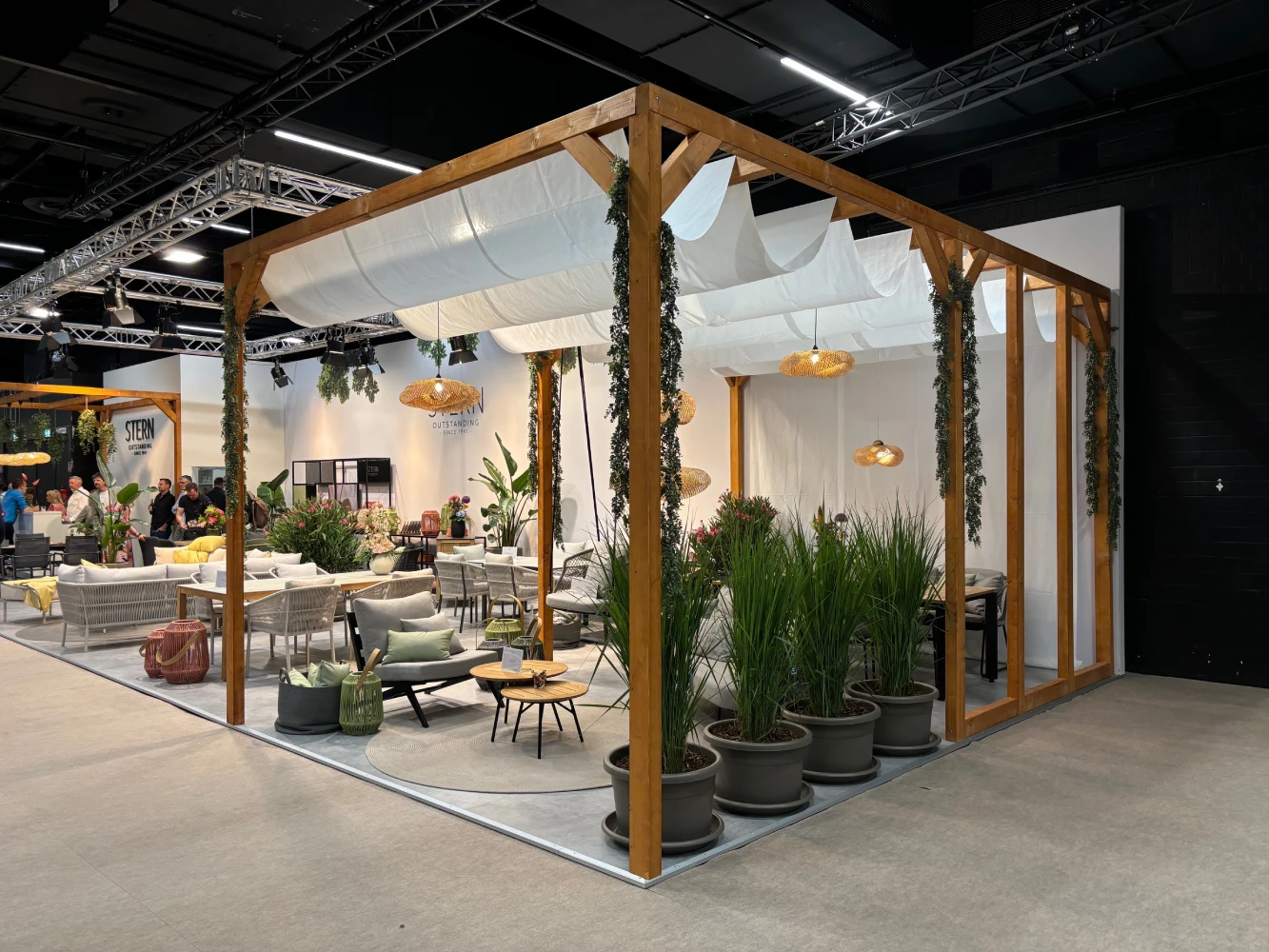 Stylish outdoor furniture display found withing global design fairs with wooden pergola and hanging lights, surrounded by potted plants. Modern, inviting, and cozy ambiance.