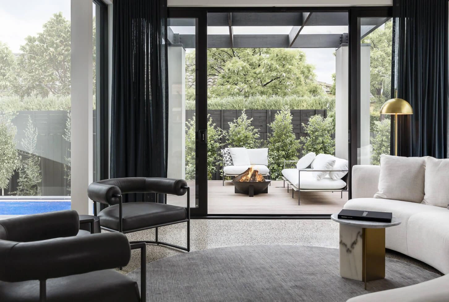 Modern living room with a view of a patio through large glass doors. Inside, a black chair, white sofa, and gold lamp; outside, white chairs and a firepit.