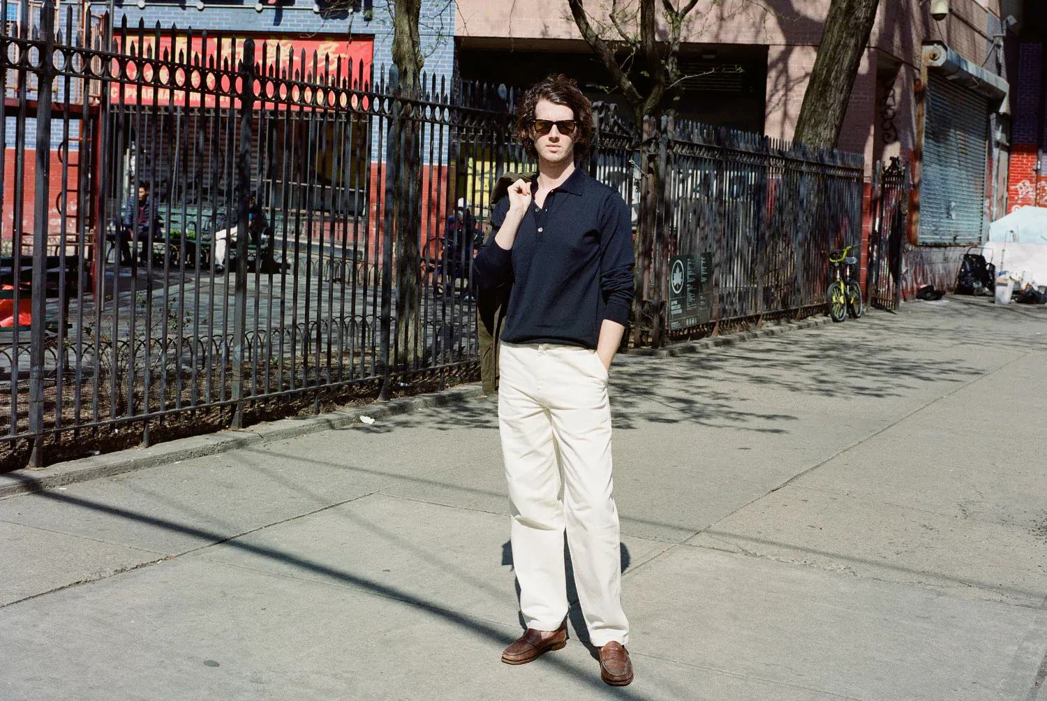 Christopher Cawley wearing sunglasses, a dark shirt, and light pants stands confidently on a sunny sidewalk. Urban setting with a fence and shop in the background.