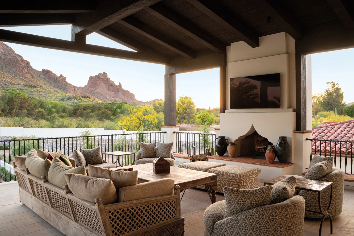 Spacious outdoor patio with cozy seating, neutral cushions, a central wooden table, fireplace, wall-mounted TV, and scenic view of desert mountains, ideal for outdoor luxury living.