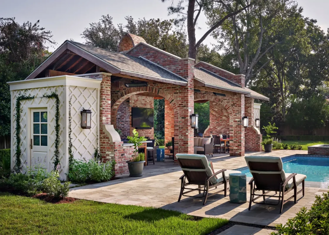 Charming outdoor space featuring a brick pavilion with arches, cozy seating, and a poolside view. Sunlit and tranquil atmosphere with lush greenery.
