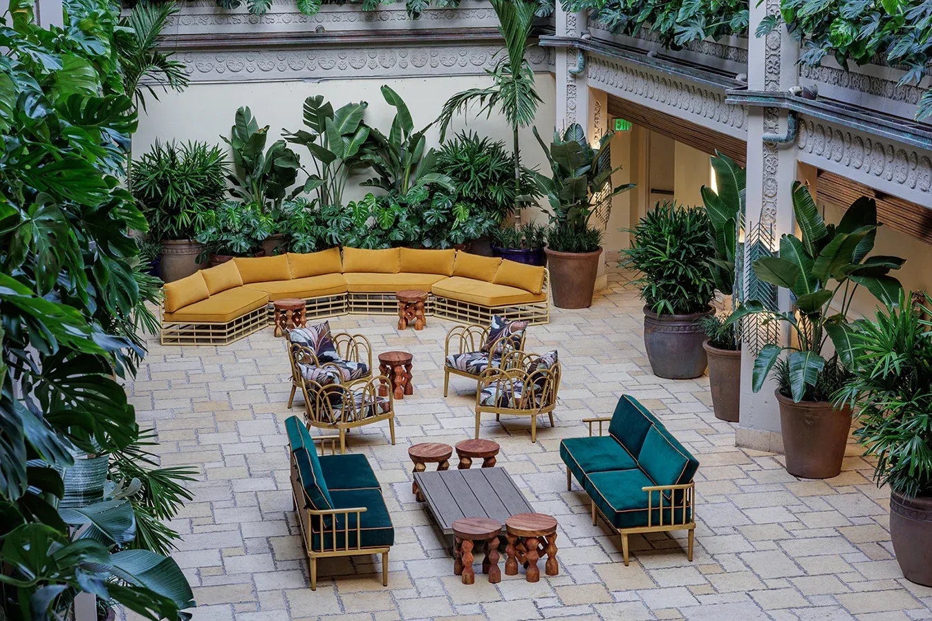 Elegant courtyard with lush greenery, yellow and green cushioned seating, wooden tables, and intricate architectural details, conveying a tranquil setting.