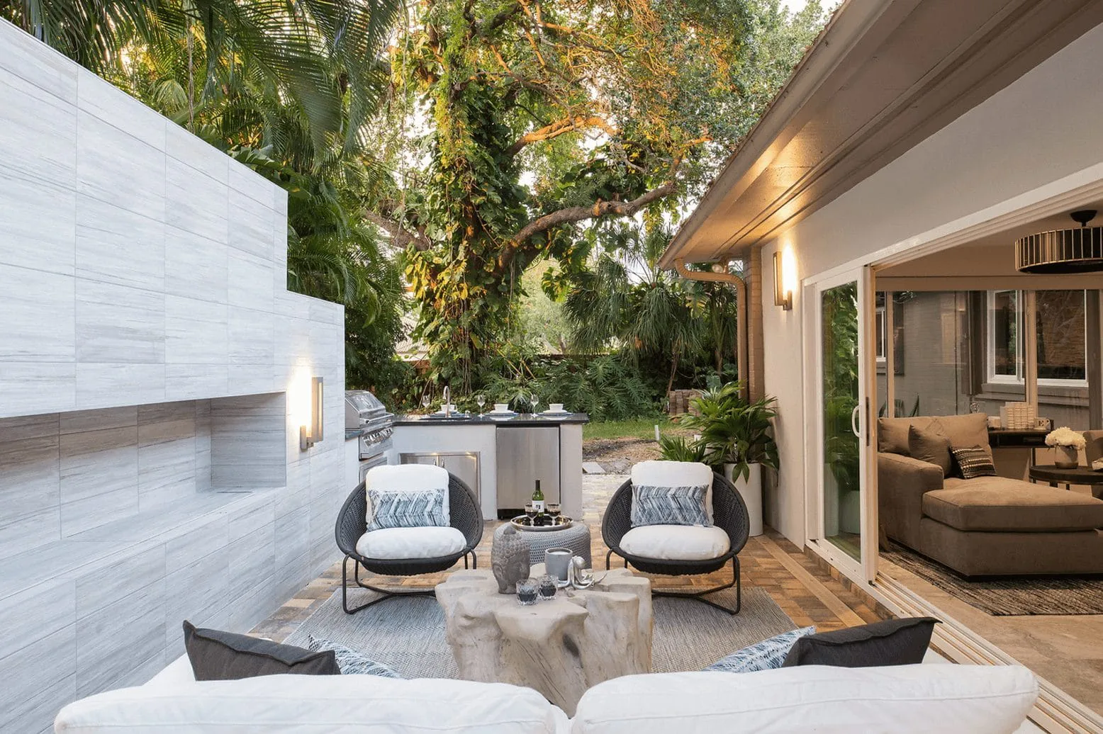 Outdoor patio featuring cushioned chairs, a rustic coffee table, and a sleek outdoor kitchen surrounded by lush greenery. Cozy and inviting ambiance.