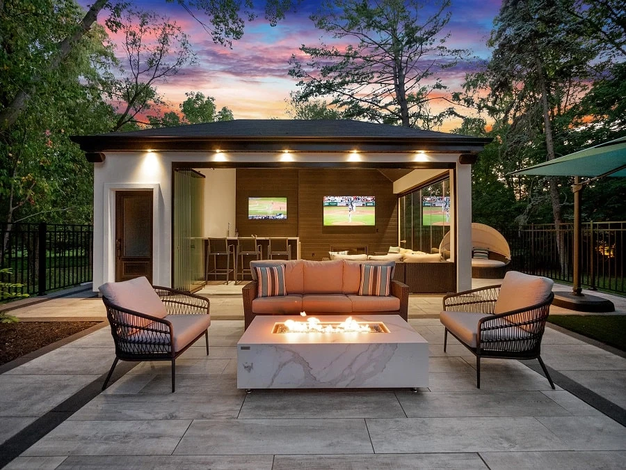 Luxurious patio with a fire pit, cozy seating, and a poolside cabana featuring TVs. The evening sky is vibrant with a colorful sunset.