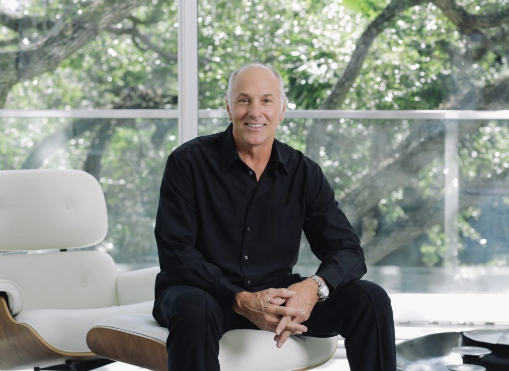 Raymond Jungles, American landscape architect and founder of Raymond Jungles Inc. in Miami, in a black shirt sits on a white chair in front of a large window, with lush green trees outside, creating a serene atmosphere.