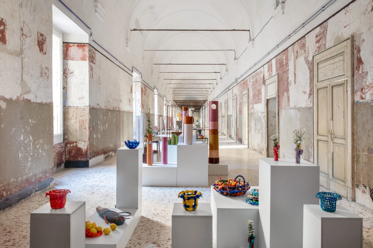 A bright gallery with high ceilings and distressed walls showcases colorful baskets and vases on white pedestals, creating a vibrant contrast.