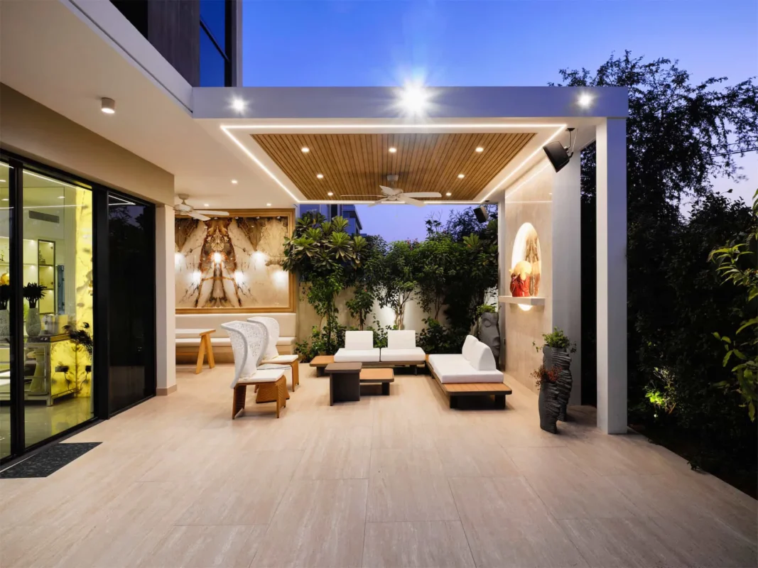Modern patio with white sofas, wooden ceiling, and plants under warm lights. Art and sculptures are displayed, creating a serene evening atmosphere.